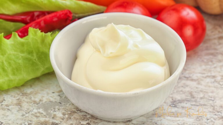 Tuisgemaakte mayonnaise in 'n glas houer, met vars bestanddele soos eiers en suurlemoensap rondom, wat 'n romerige en smaakvolle toevoeging tot etes aandui